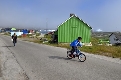 Greenland, west coast, Disko Island, houses in the village of Qeqertarsuaq and icebergs in the background, child on bicycle