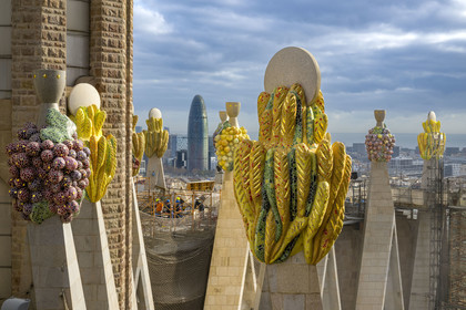 Spain, Catalonia, Barcelona, Eixample district, Sagrada Familia basilica by Catalan modernist architect Antoni Gaudi, listed as a UNESCO World Heritage Site, peaks topped with mosaics in the shape of fruits surrounding the construction site on the roof of the nave at the rear of the future facade of the Glory, the Torre Agbar by architect Jean Nouvel in the background