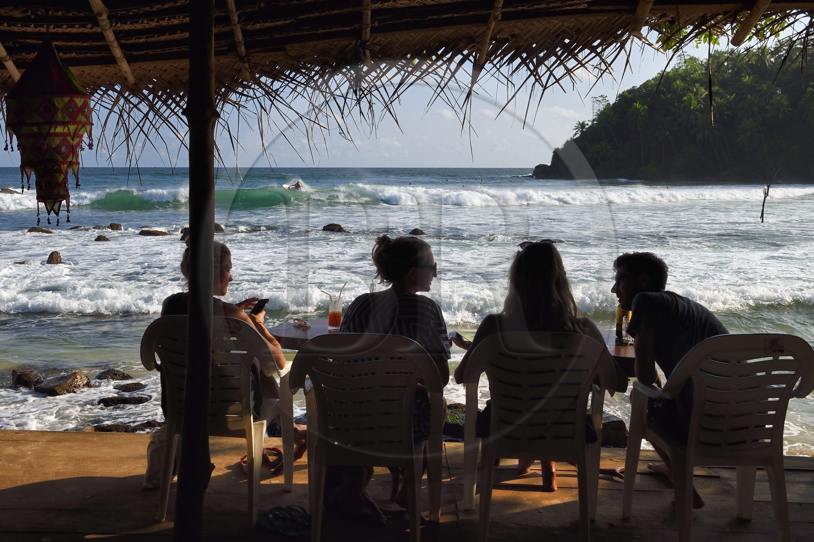 Sri Lanka, Province du Sud, Matara (district), Weligama, plage de Mirissa, jeunes gens attablés dans un restaurant de plage observant un surfeur