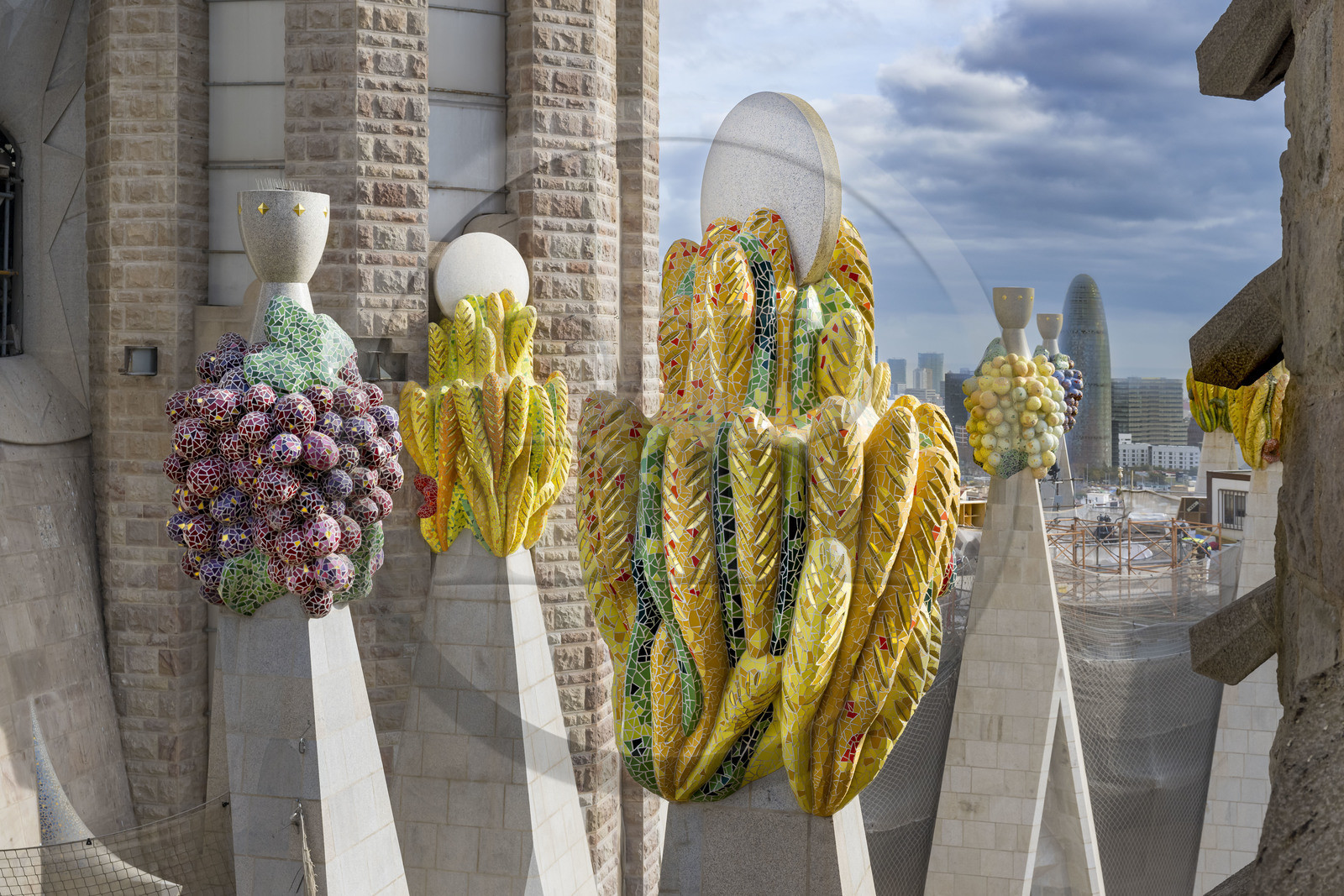 Spain, Catalonia, Barcelona, Eixample district, Sagrada Familia basilica by Catalan modernist architect Antoni Gaudi, listed as a UNESCO World Heritage Site, peaks topped with mosaics in the shape of fruits, the Torre Agbar by architect Jean Nouvel in the background