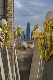 Spain, Catalonia, Barcelona, Eixample district, Sagrada Familia basilica by Catalan modernist architect Antoni Gaudi, listed as a UNESCO World Heritage Site, peaks topped with mosaics in the shape of fruits surrounding the construction site on the roof of the nave at the rear of the future facade of the Glory, the Torre Agbar by architect Jean Nouvel in the background