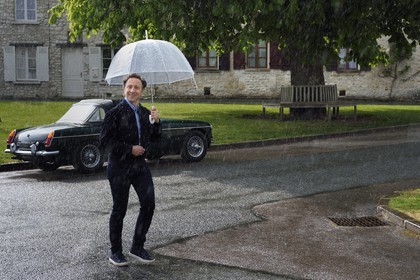 France, Yvelines (78), Montchauvet, tournage pour la télévision du Village Préféré des Français avec Stéphane Bern, Stéphane Bern sous la pluie sur la place de l'église