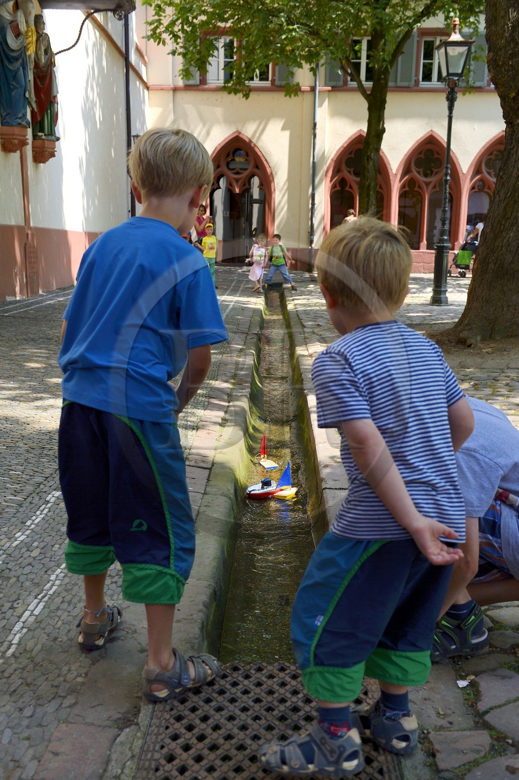 Germany, Baden-Wurttemberg, Freiburg im Breisgau, Rathausplatz, children playing with small boats in one of the Bächle which are small open channel lining the sidewalks