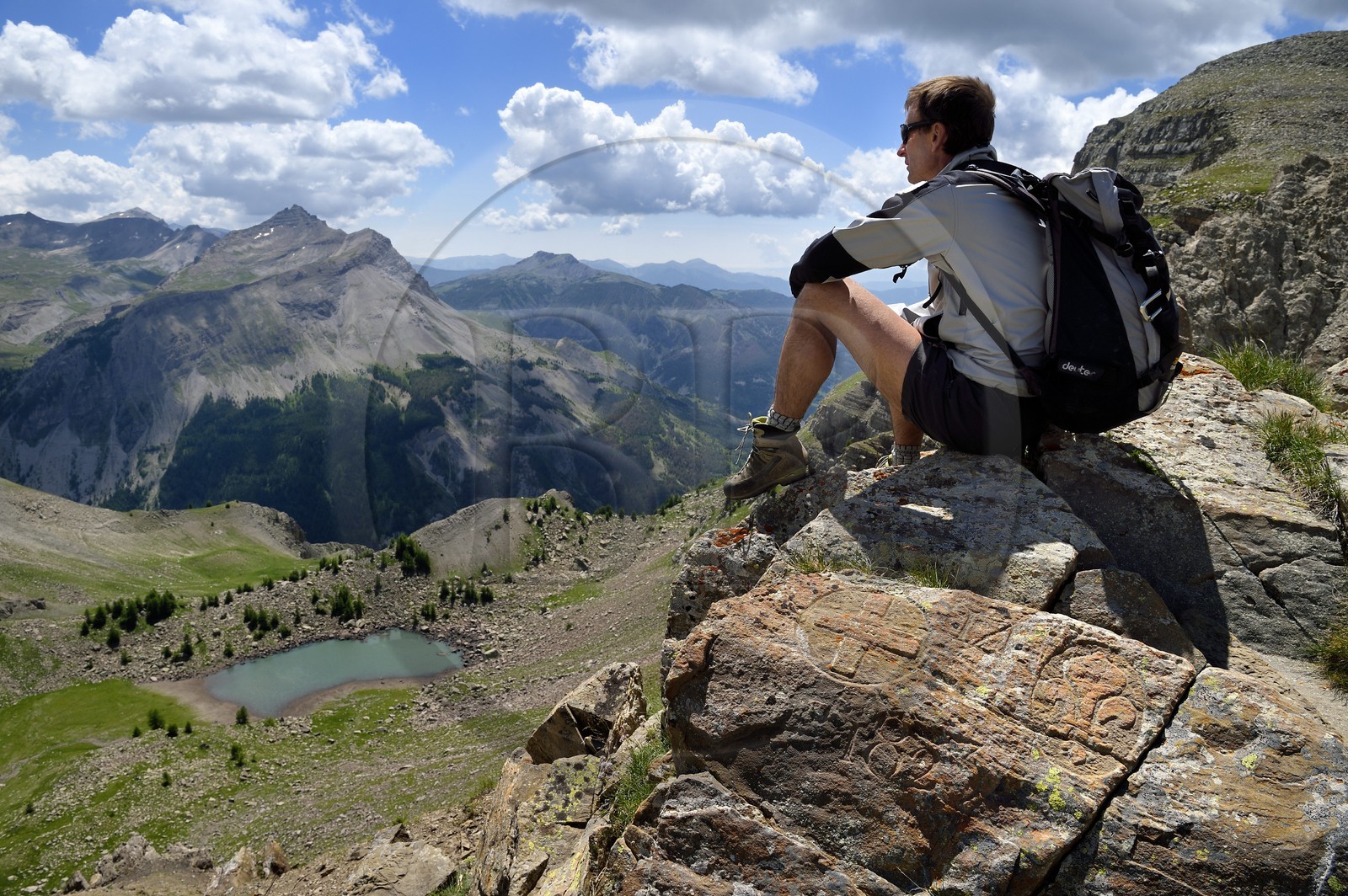 France, Alpes-de-Haute-Provence (04), Uvernet-Fours, parc national du Mercantour, vallée de l'Ubaye, sentier de randonnée du circuit des lacs du col de la Cayolle au Pas du Lausson, François Breton garde moniteur du parc assis sur une borne gravée sur les rochers délimitant la frontière entre le duché de Savoie et le comté de Nice, la vallée du Haut-Var et lac du Lausson (Alpes-Maritimes) en arrière plan