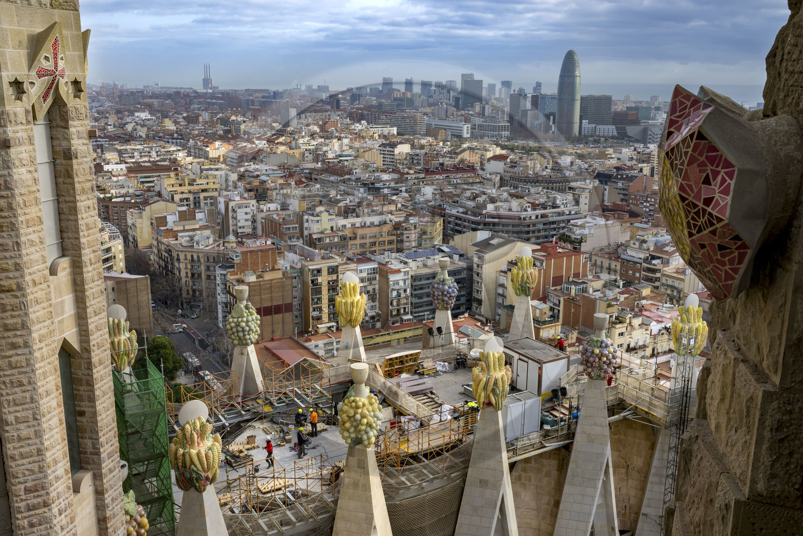 Spain, Catalonia, Barcelona, Eixample district, Sagrada Familia basilica by Catalan modernist architect Antoni Gaudi, listed as a UNESCO World Heritage Site, peaks topped with mosaics in the shape of fruits surrounding the construction site on the roof of the nave at the rear of the future facade of the Glory, the Torre Agbar by architect Jean Nouvel in the background