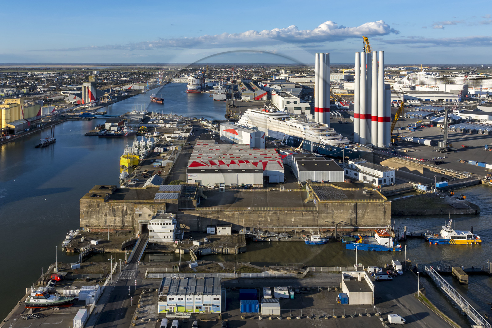 France, Loire-Atlantique (44), Saint-Nazaire, l’écluse Est et l'écluse fortifiée de l'ancienne base sous-marine allemande construite lors de la dernière guerre mondiale au premier plan et le chantier du super-yacht de luxe Ritz-Carlton Luminara dans la forme Joubert, les tours d'éoliennes à droite sont stockées avant embarquement, le paquebot de 333m MSC World America construit par les Chantiers de l'Atlantique en arrière plan (vue aérienne)